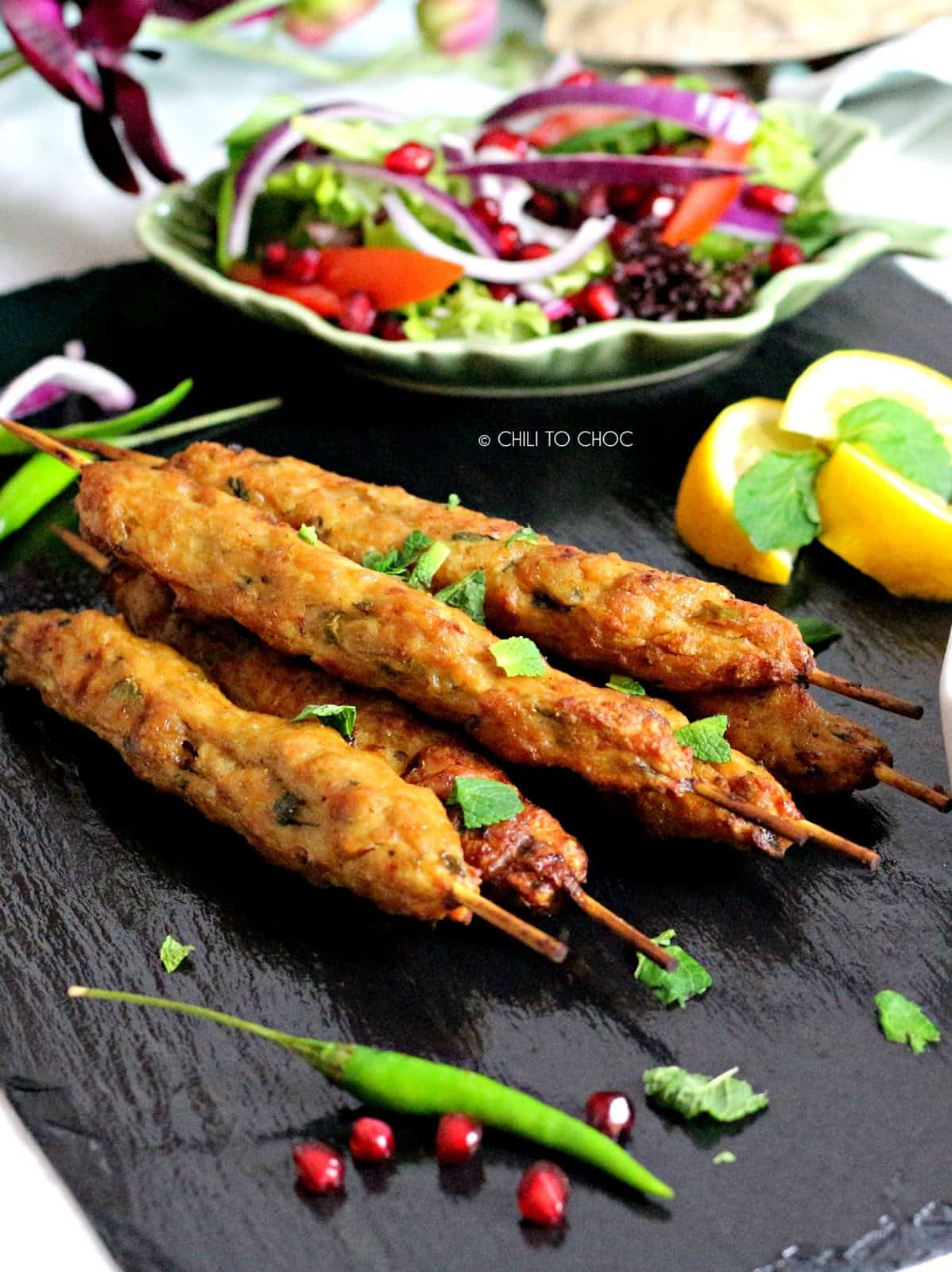 Skewers of ground beef stacked on a black board with a salad plate behind them.