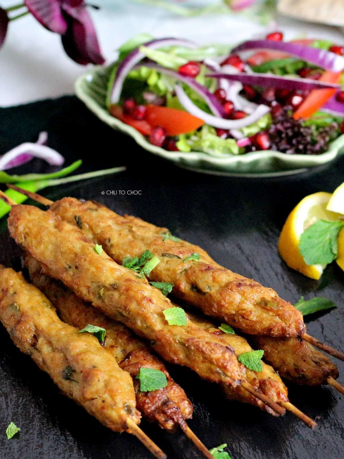 Closeup of ground beef skewers with a mixed salad behind it and lemon wedges on the side.