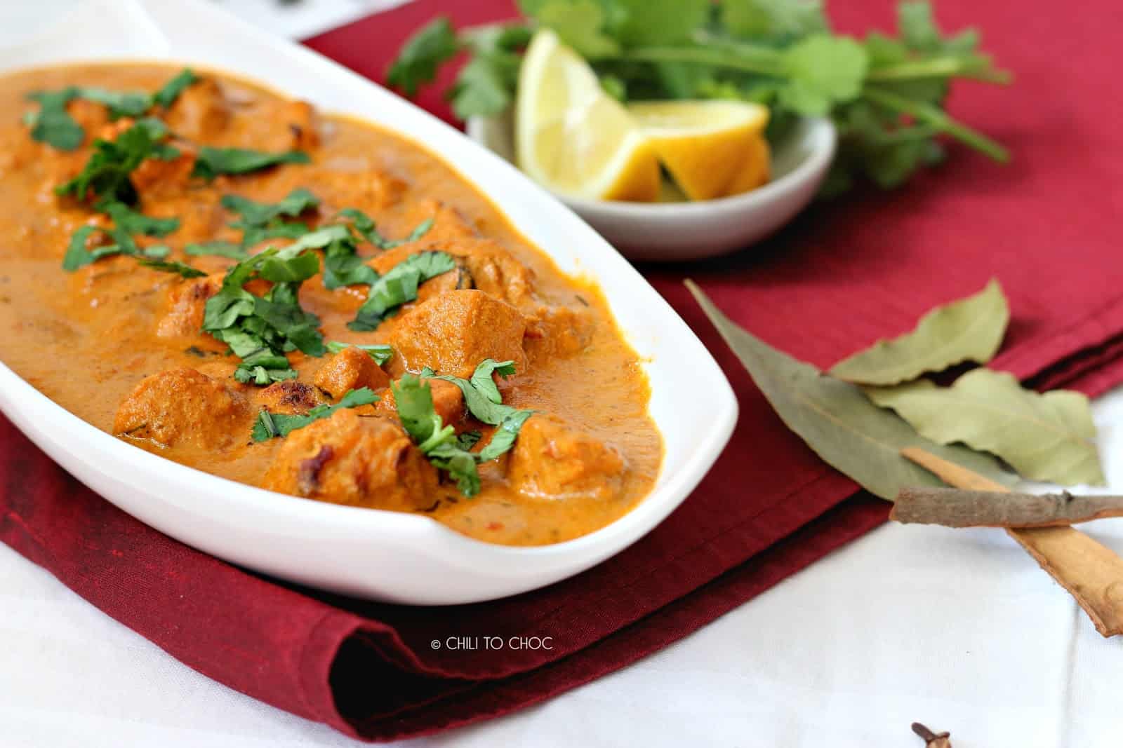 Chicken tikka masala in a white dish with bay leaves on the side and garnishes at the back.