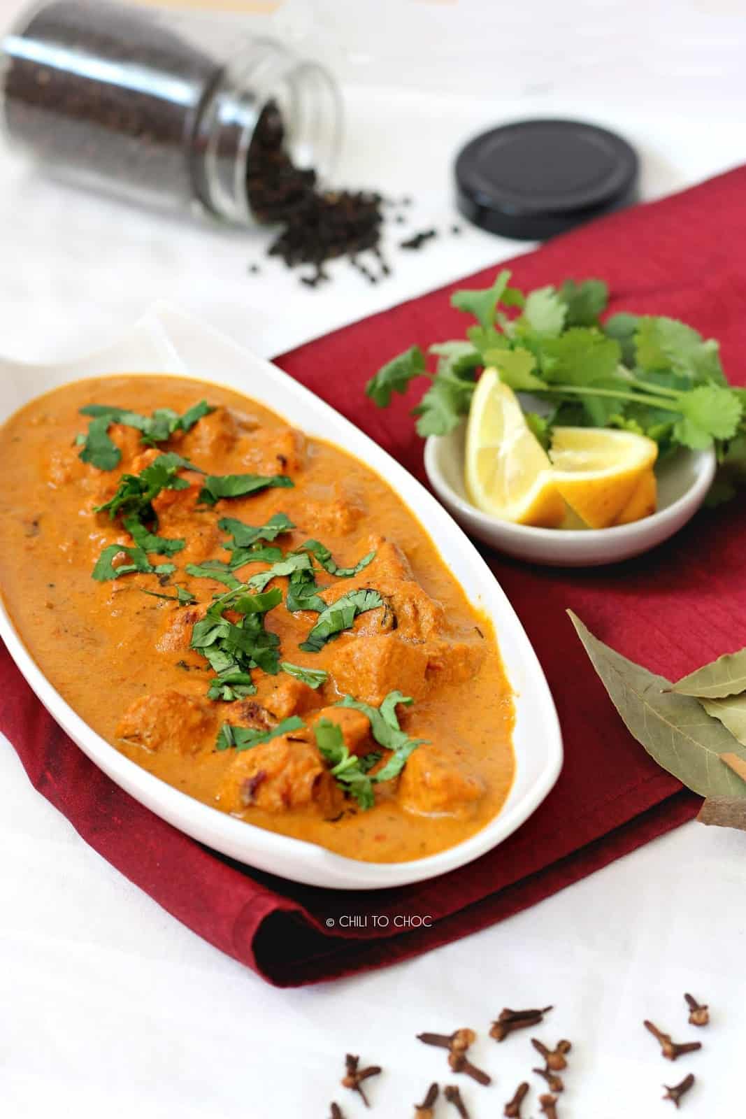 Chicken tikka masala in a white dish on a red piece of fabric with garnish at the back.