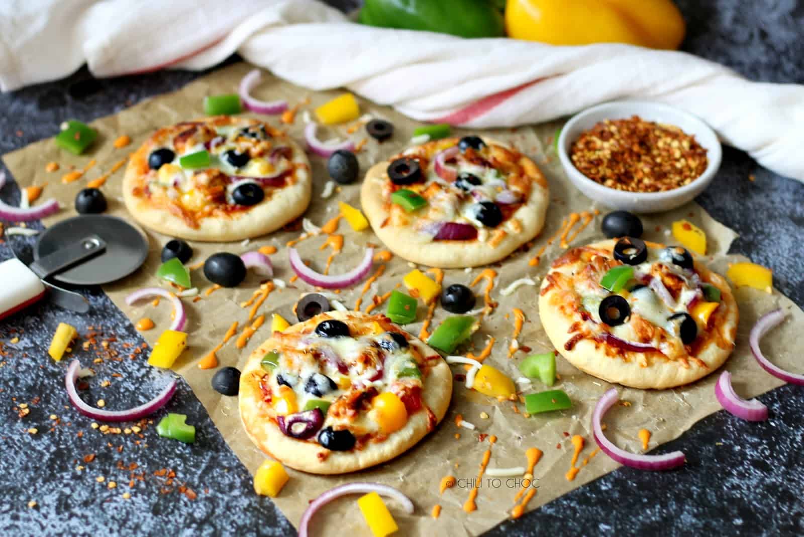 Mini pizzas on a parchment paper surrounded by their toppings.