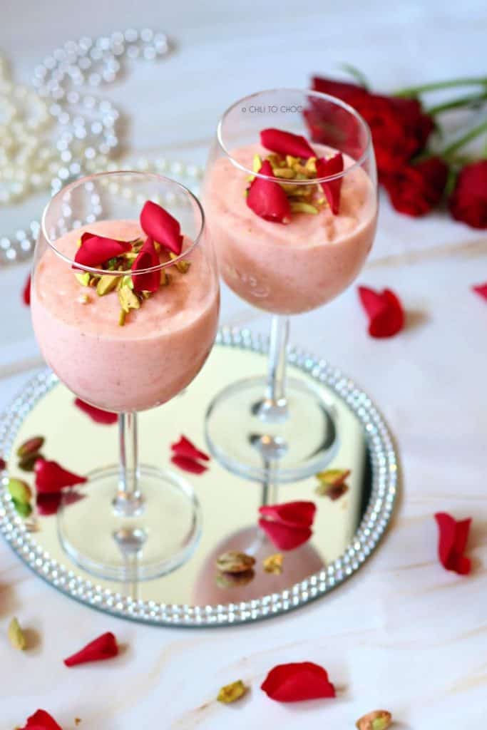 Two firni in stem glasses on a mirror plate decorated with rose petals and chopped pistachio