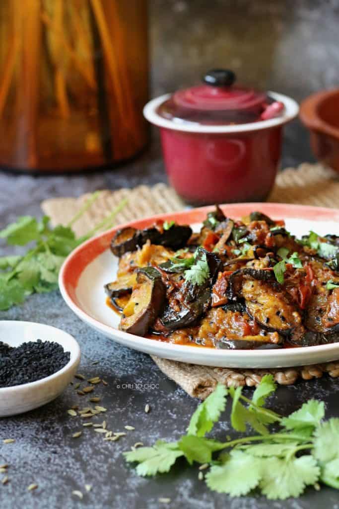 Eggplant curry in a plate with nigella seeds in front and fennel seeds on the backdrop