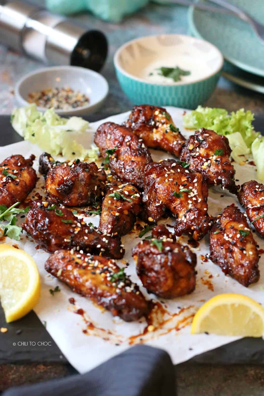 Sticky wings on a parchment paper surrounded by its dip, toasted sesame seeds and lemon wedges.