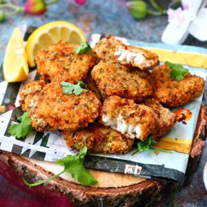 spicy fish nuggets on a piece of paper garnished with chopped coriander and lemon wedges