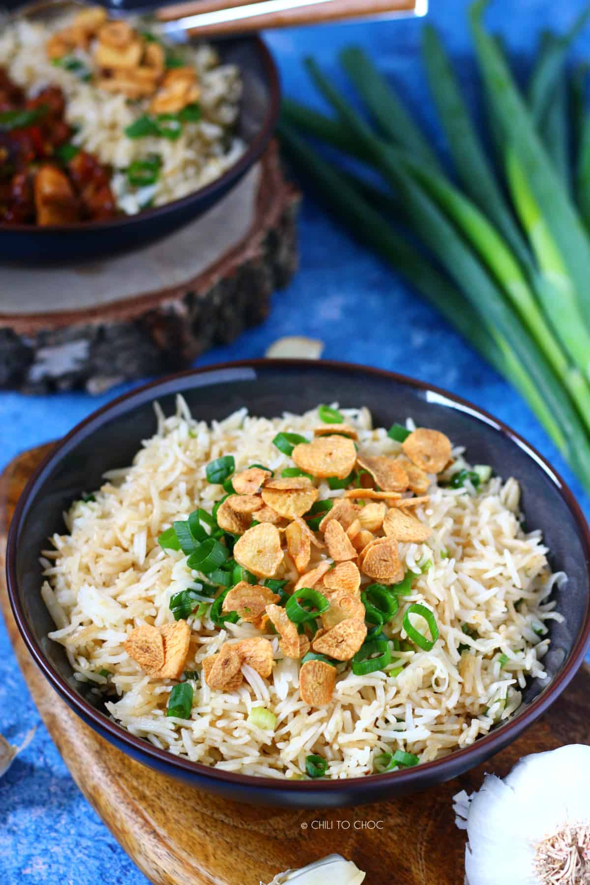 Garlic butter fried rice topped with garlic chips and spring onion in a shallow dish with a bowl of Chinese chili chicken and garlic rice in the back