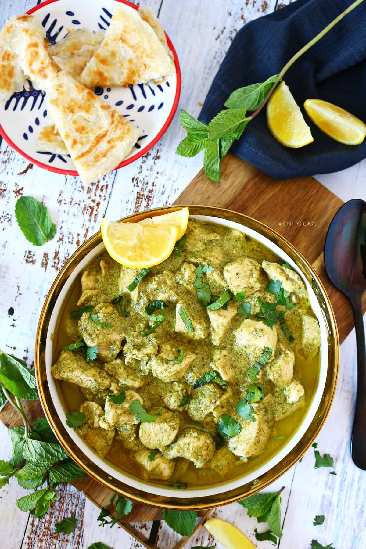 Hariyali chicken bowl on a wooden board with spoon, mint, lemon wedges and parathas around it. 