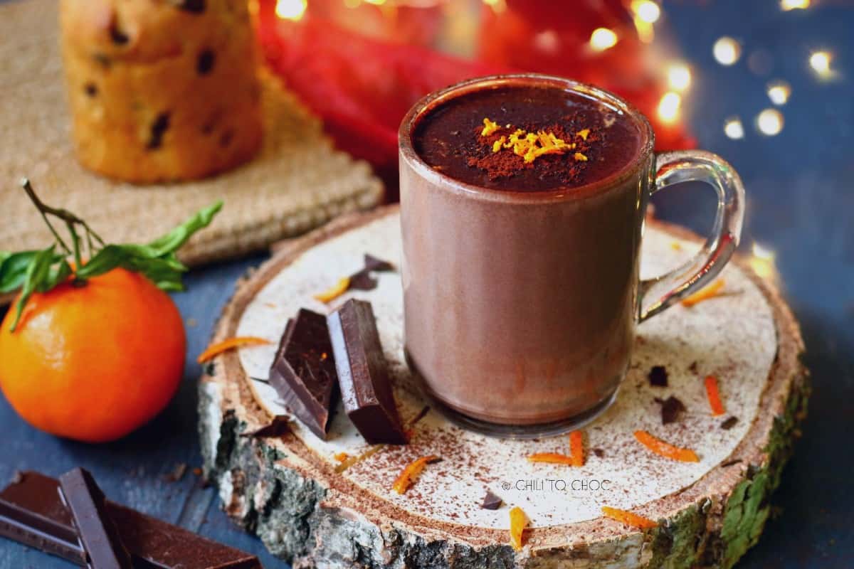 A mug of orange hot chocolate with boke lights an muffin in the background.