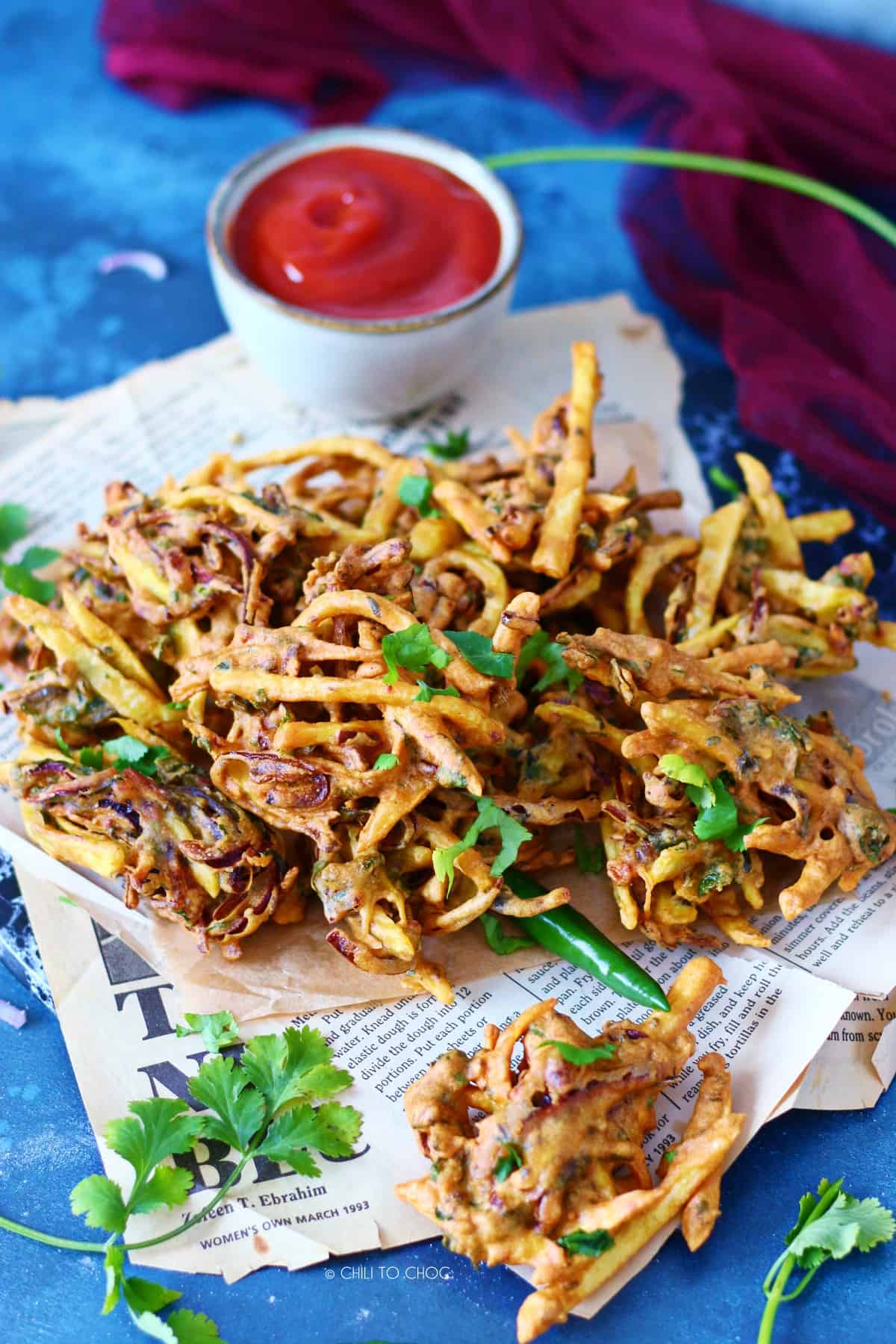 Pakoras on a magazine paper garnished with fresh coriander and green chilies with a ketchup bowl on the side.