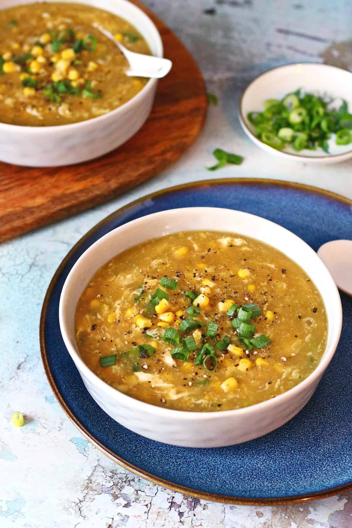 Two bowls of corn soup, one on a blue plate and the second on a wooden slab.