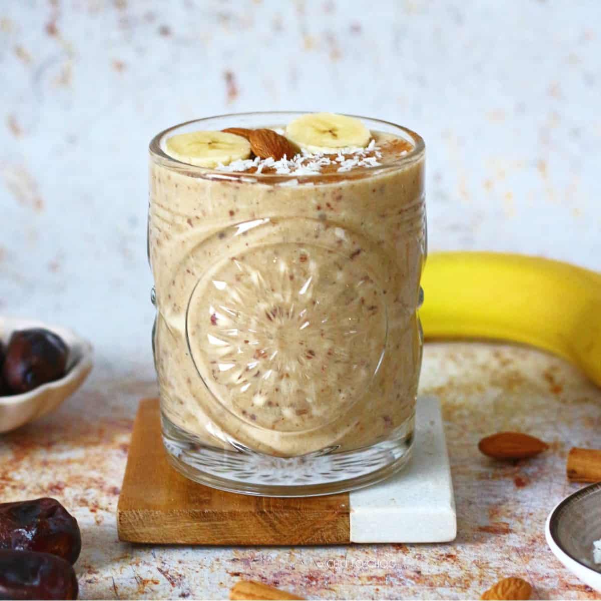 Banana Date Smoothie in a glass, on a coaster with a banana and dates around it.