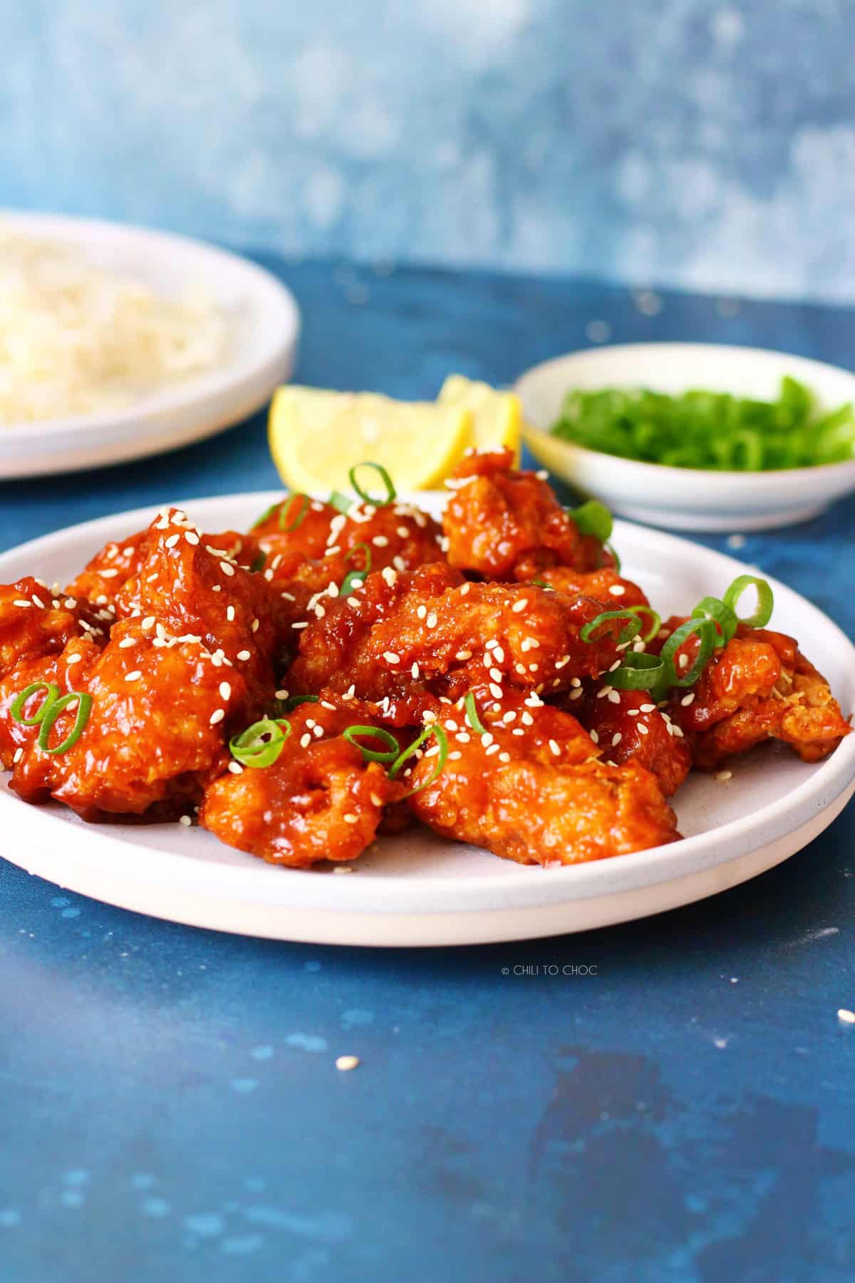 Honey sriracha chicken garnished with sesame seeds and green onion with rice and some ingredients at the back.