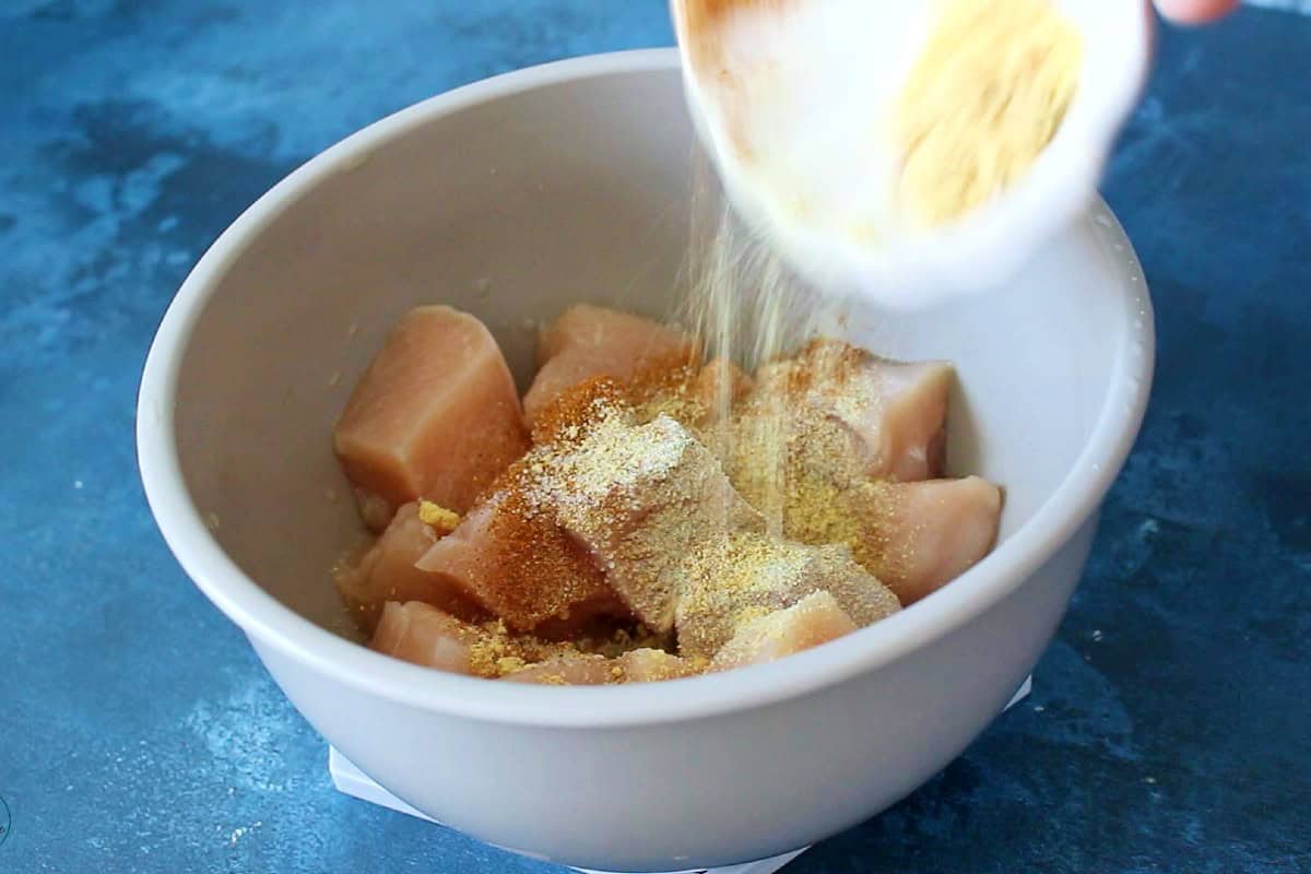 Adding seasoning to raw chicken pieces in a bowl.