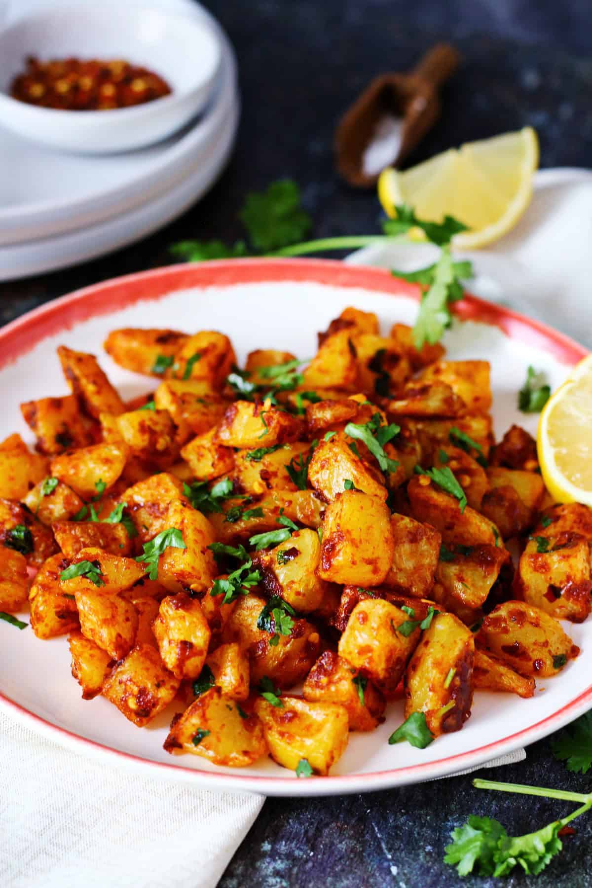 A plate of Batata Harra with lemon wedges, serving plates and fresh cilantro stem at the side.