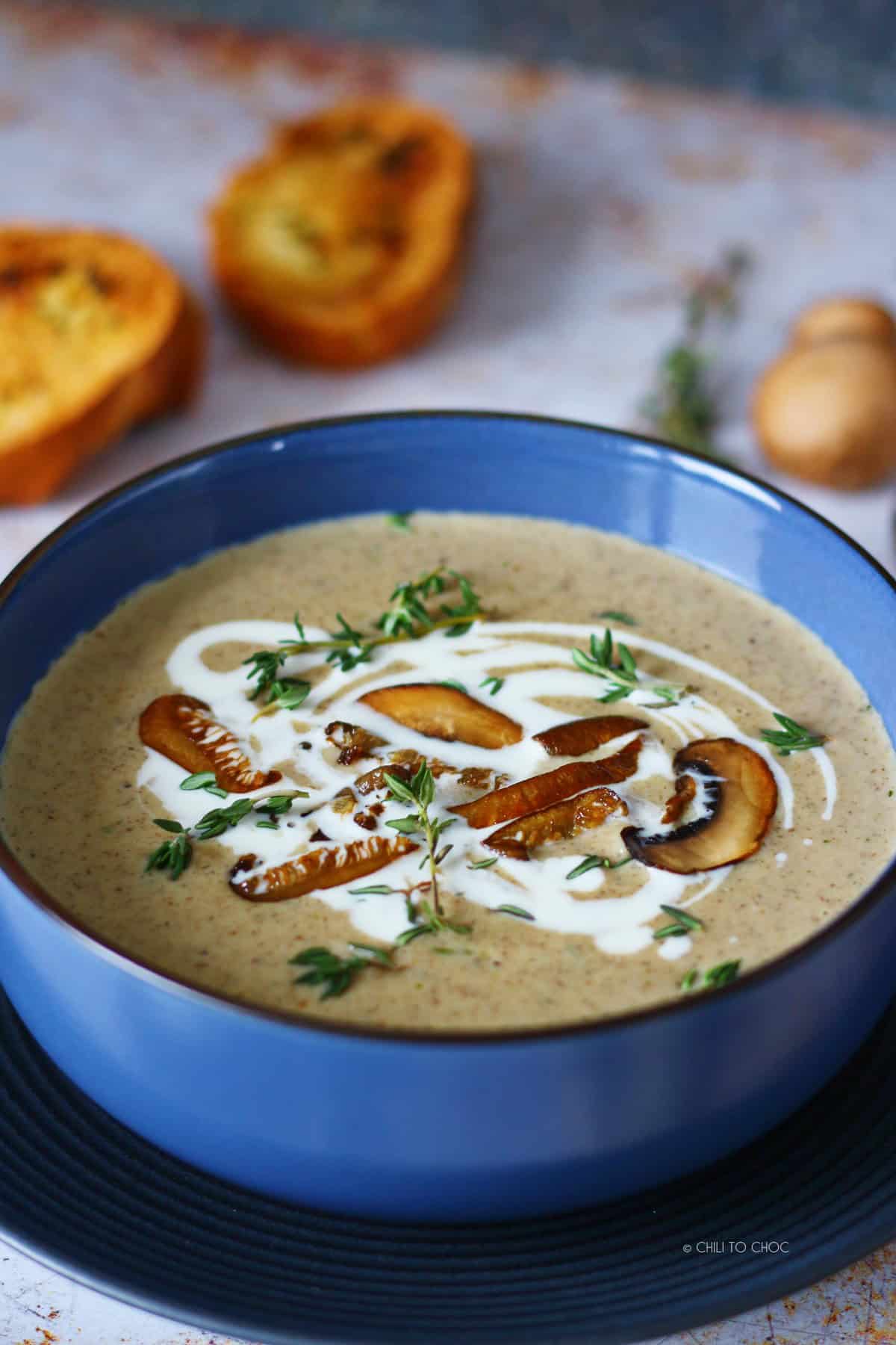 Garnished mushroom soup in a blue bowl with garlic bread at the back.