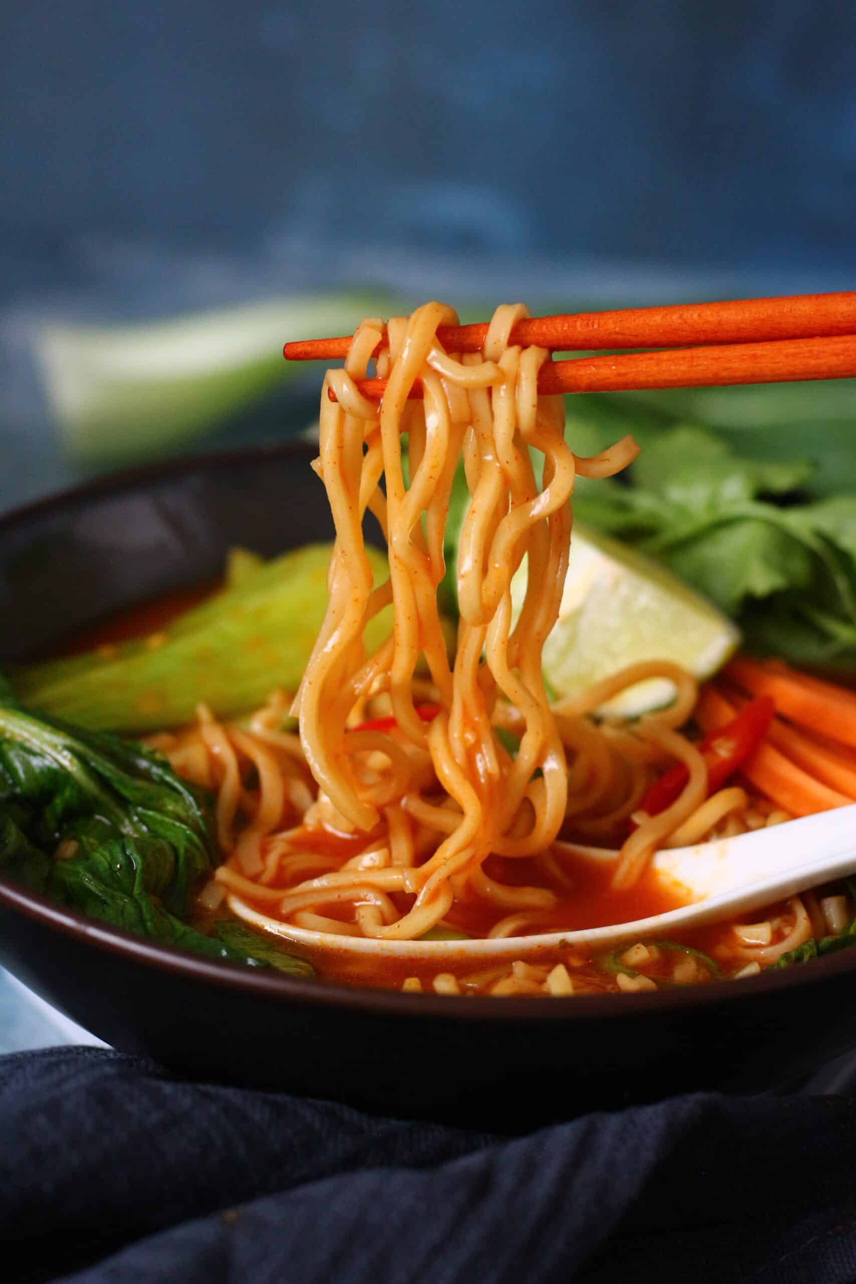 A bowl of soup with noodles held by chopsticks above the bowl. It includes leafy greens and a wedge of lime in a red broth.