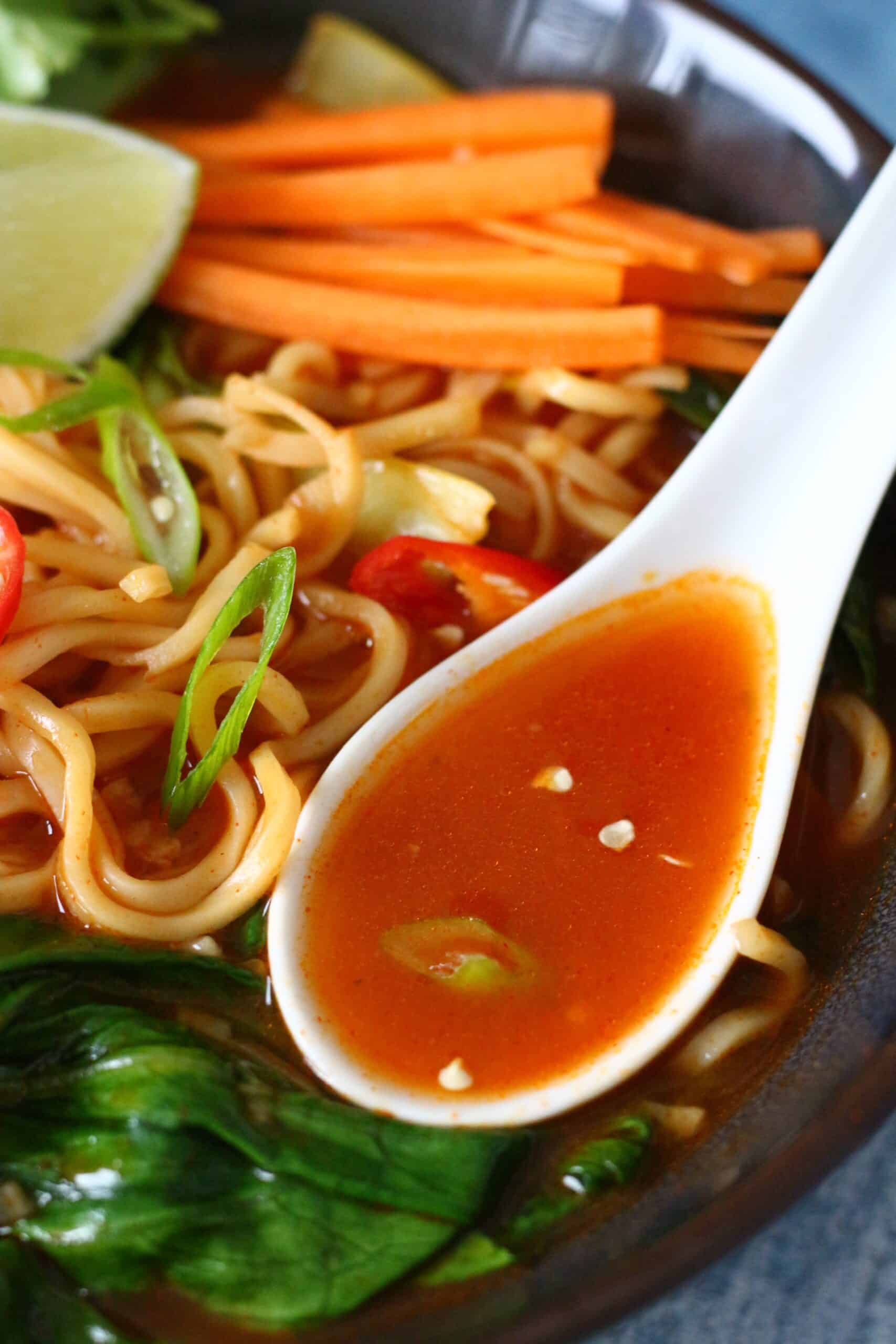 A bowl of Asian noodle soup with orange broth in a white soup spoon.