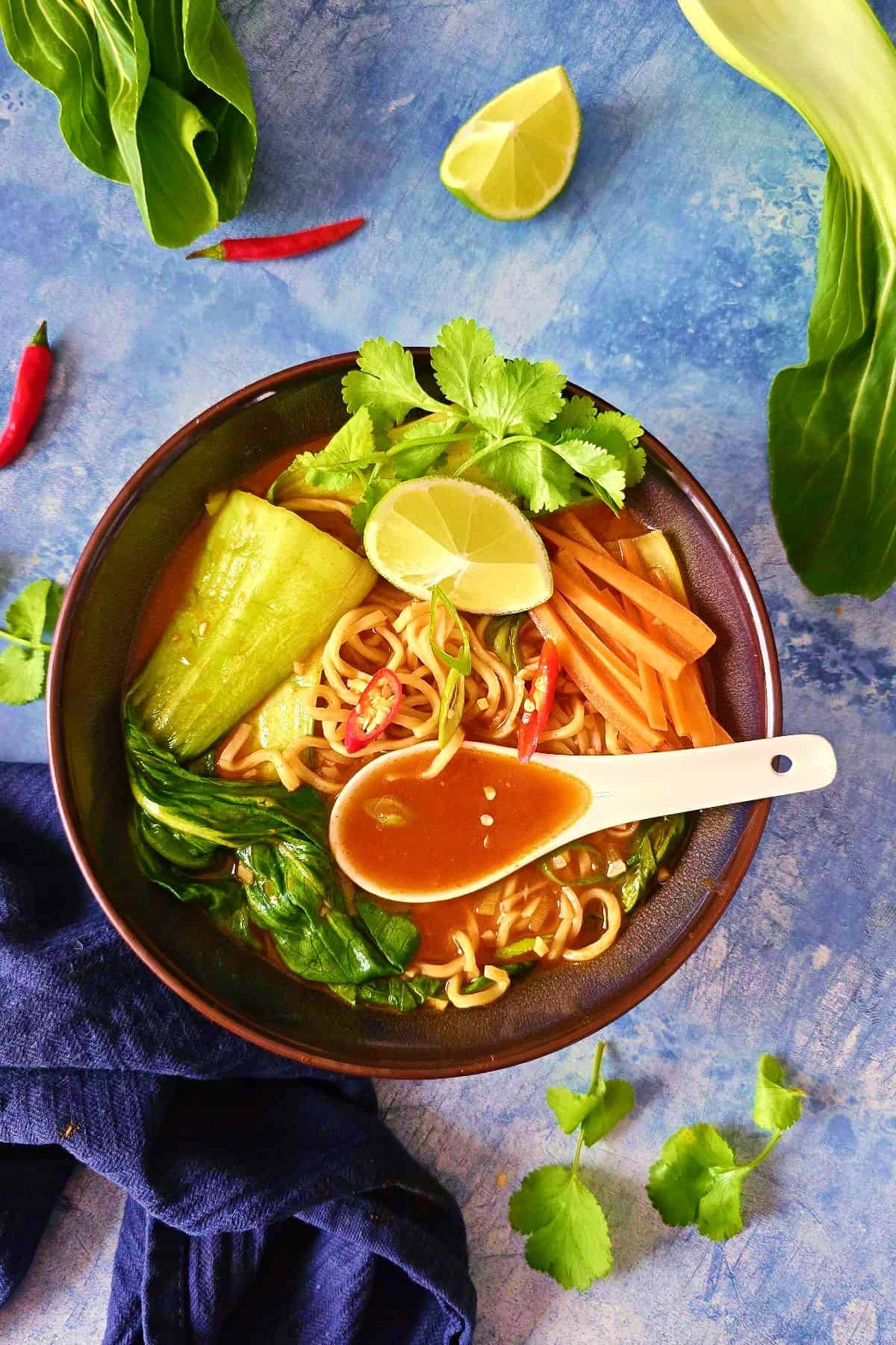 A bowl of Asian noodle soup with orange broth, bok choy, carrots, lime wedges, cilantro, and a white spoon on a blue background.