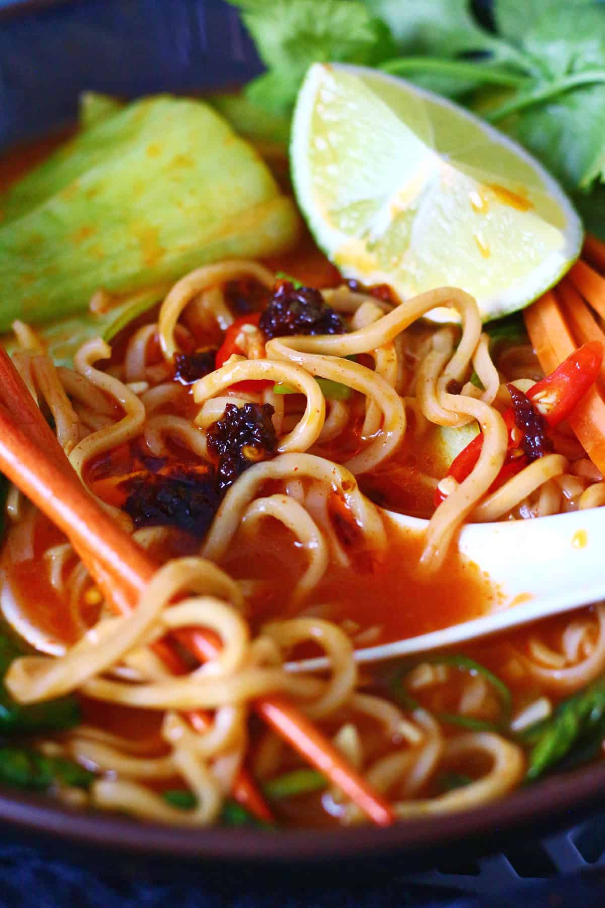 Close-up of spicy noodle soup with chili oil, bok choy, lime, spoon and chopsticks.