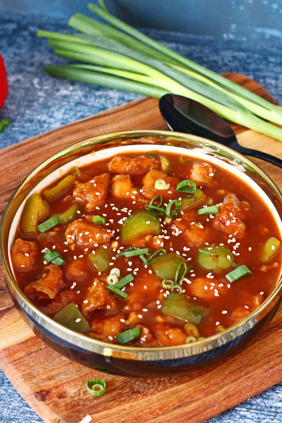 A dish of Chicken Manchurian on a wooden board with a serving spoon and green onions at the back.