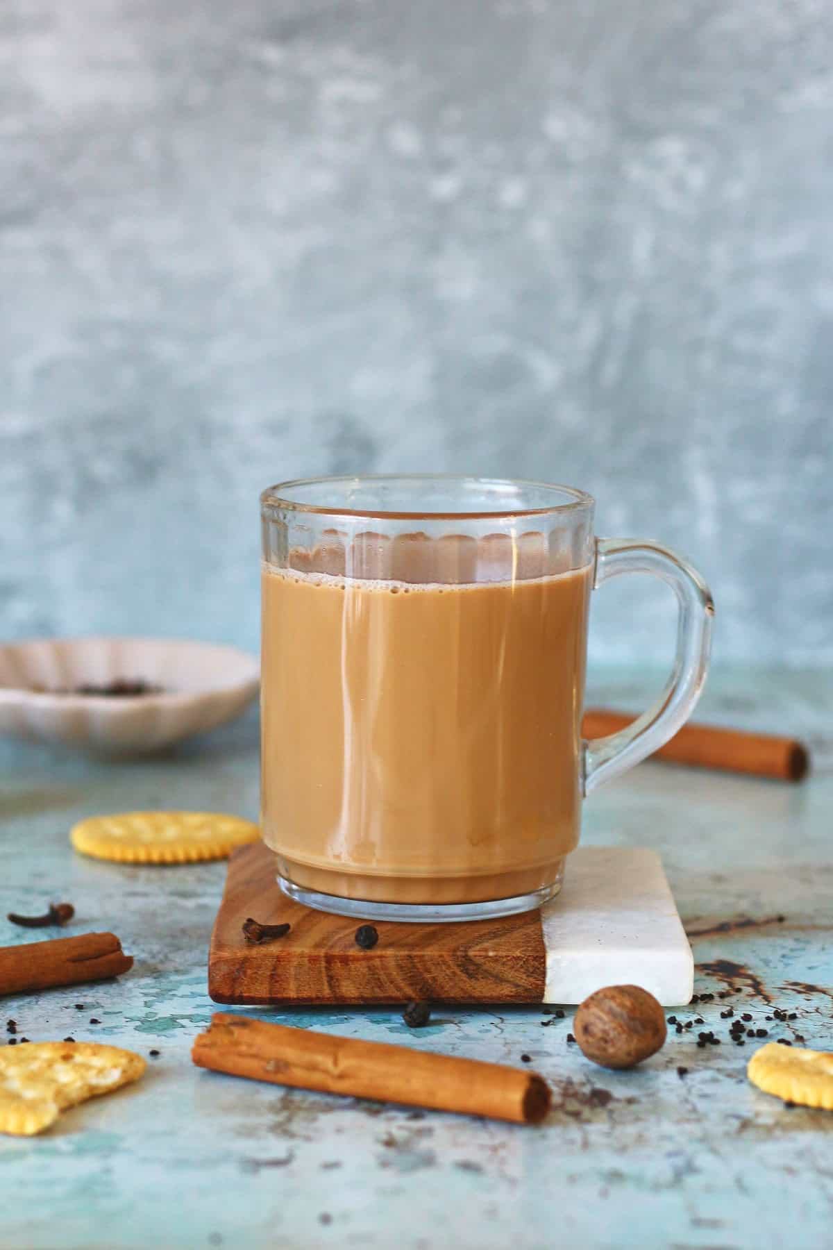 A cup of masala chai on a coaster with whole spices and cookies around it.