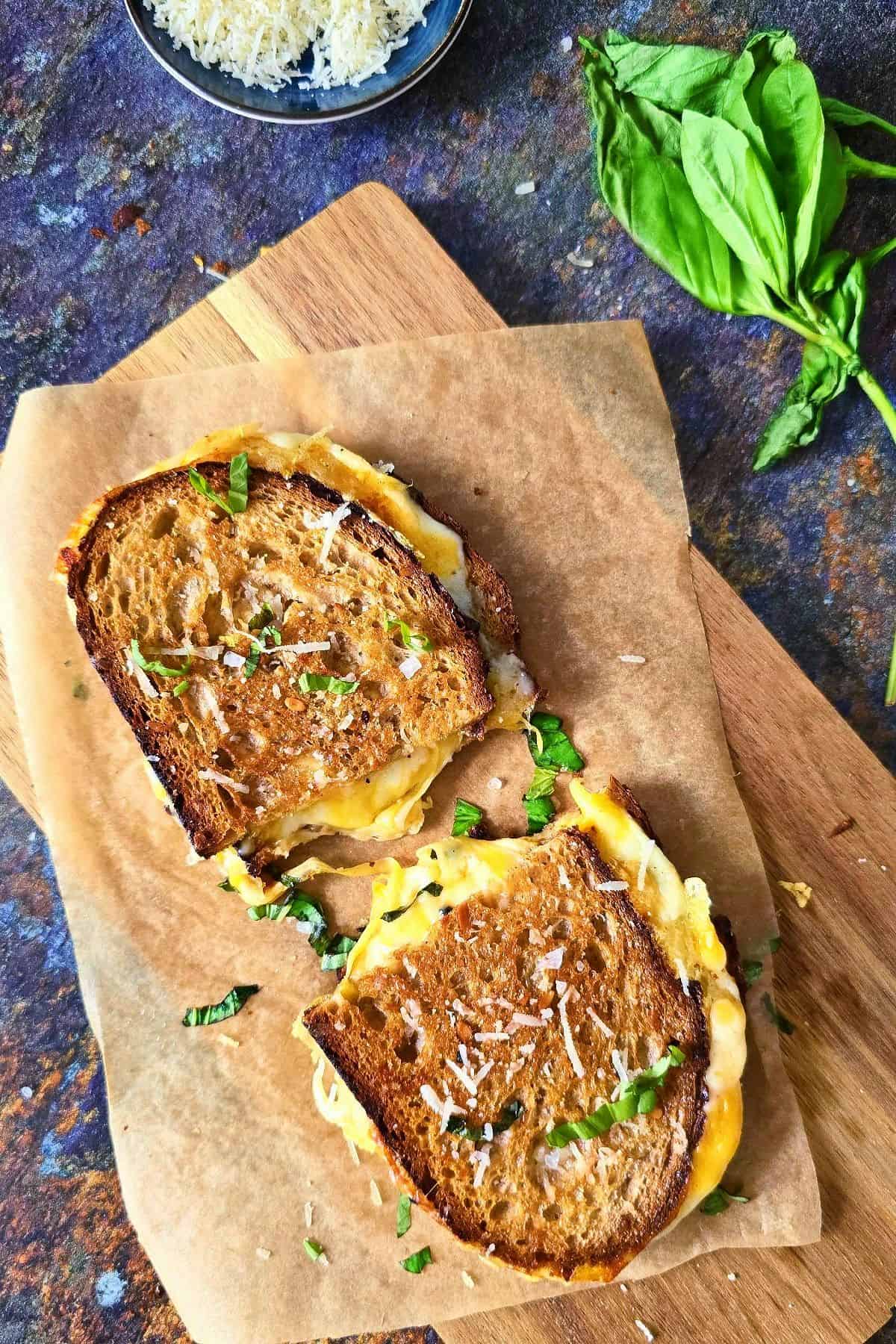 A grilled cheese sandwich halved and placed on parchment paper with fresh basil on the side.