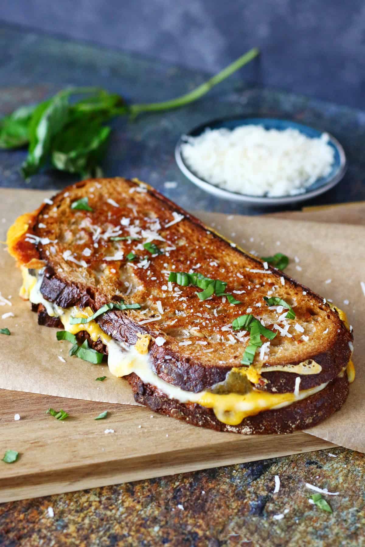 A sourdough grilled cheese sandwich on a parchment paper garnished with chopped basil and grated Parmesan.