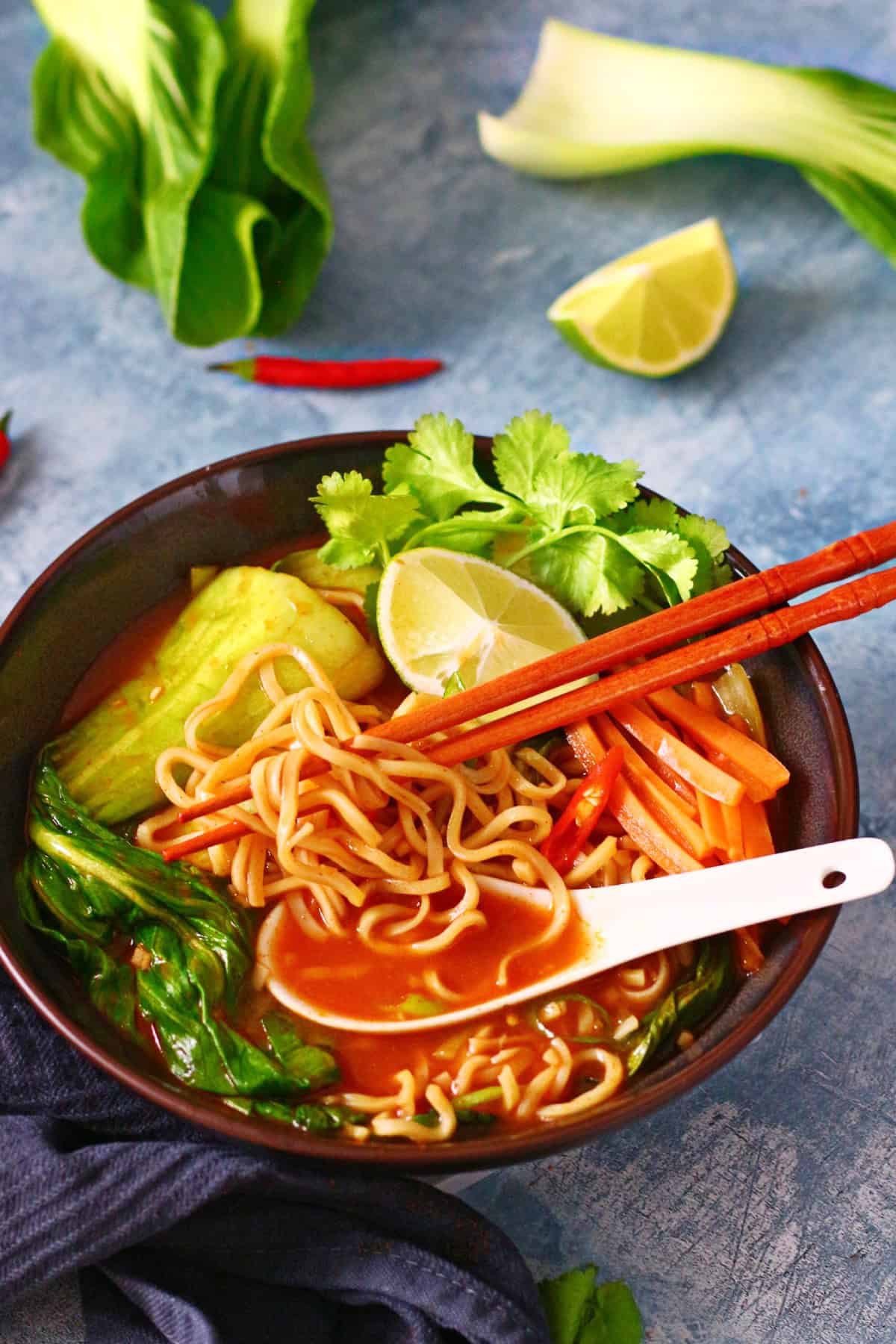 A bowl of Asian noodle soup with bok choy, carrot slices, lime wedges, and cilantro, accompanied by chopsticks and a spoon.