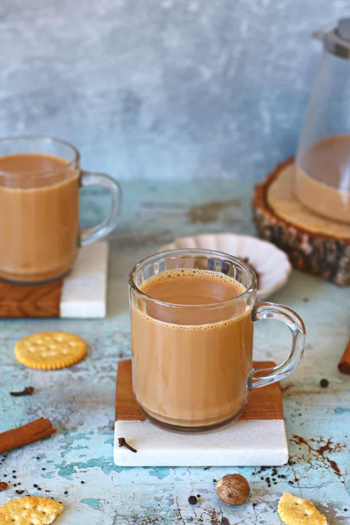 Two cups of masala chai on coasters with whole spices and a cookie around it.