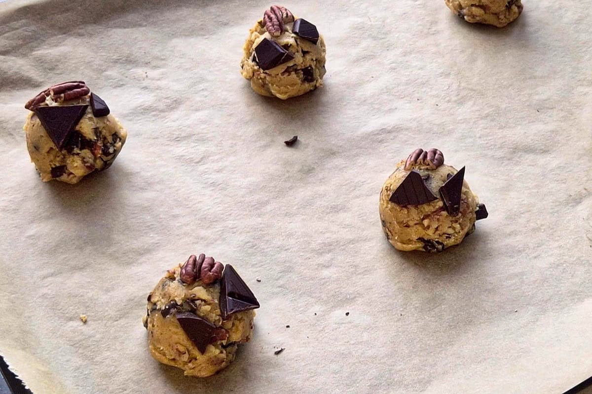 Cookie balls on a baking tray topped with pecan and chopped chocolate.