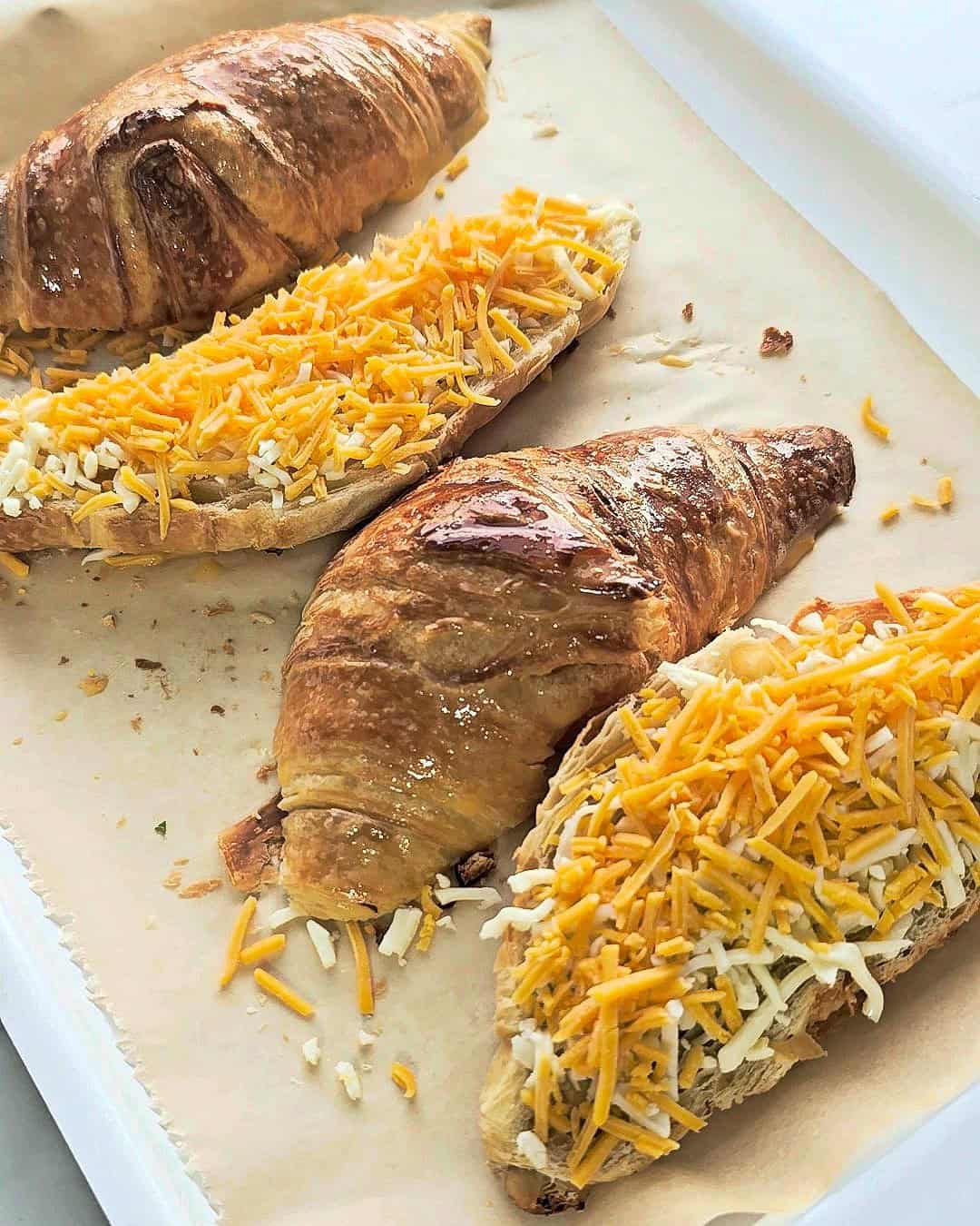 Sliced croissants on a baking tray with their bottoms covered with shredded cheese.