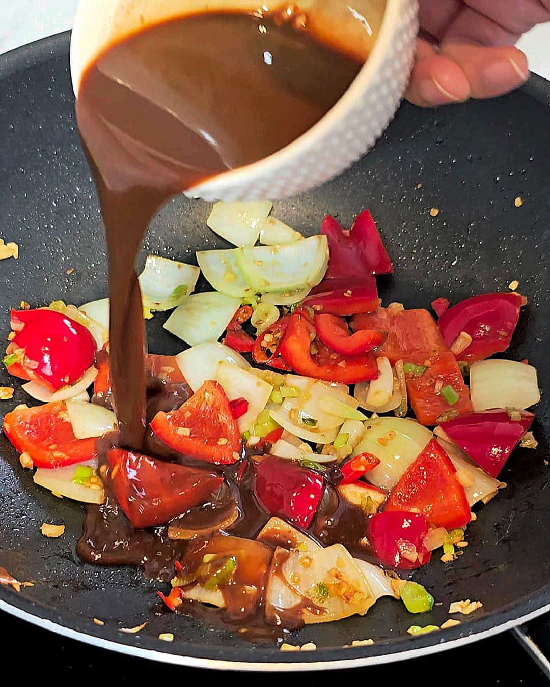 Pouring sauce from a bowl into a wok with sauteed onions and peppers.