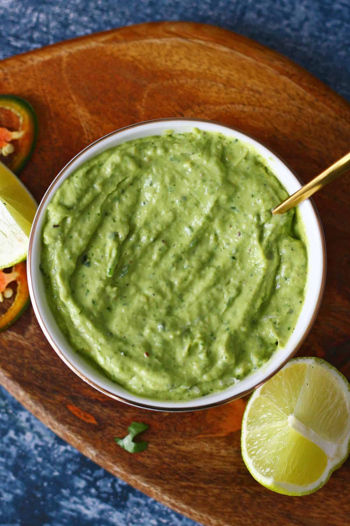 Overhead shot of avocado crema in a white bowl with spoon and lime wedges and jalapeno slices on the side.