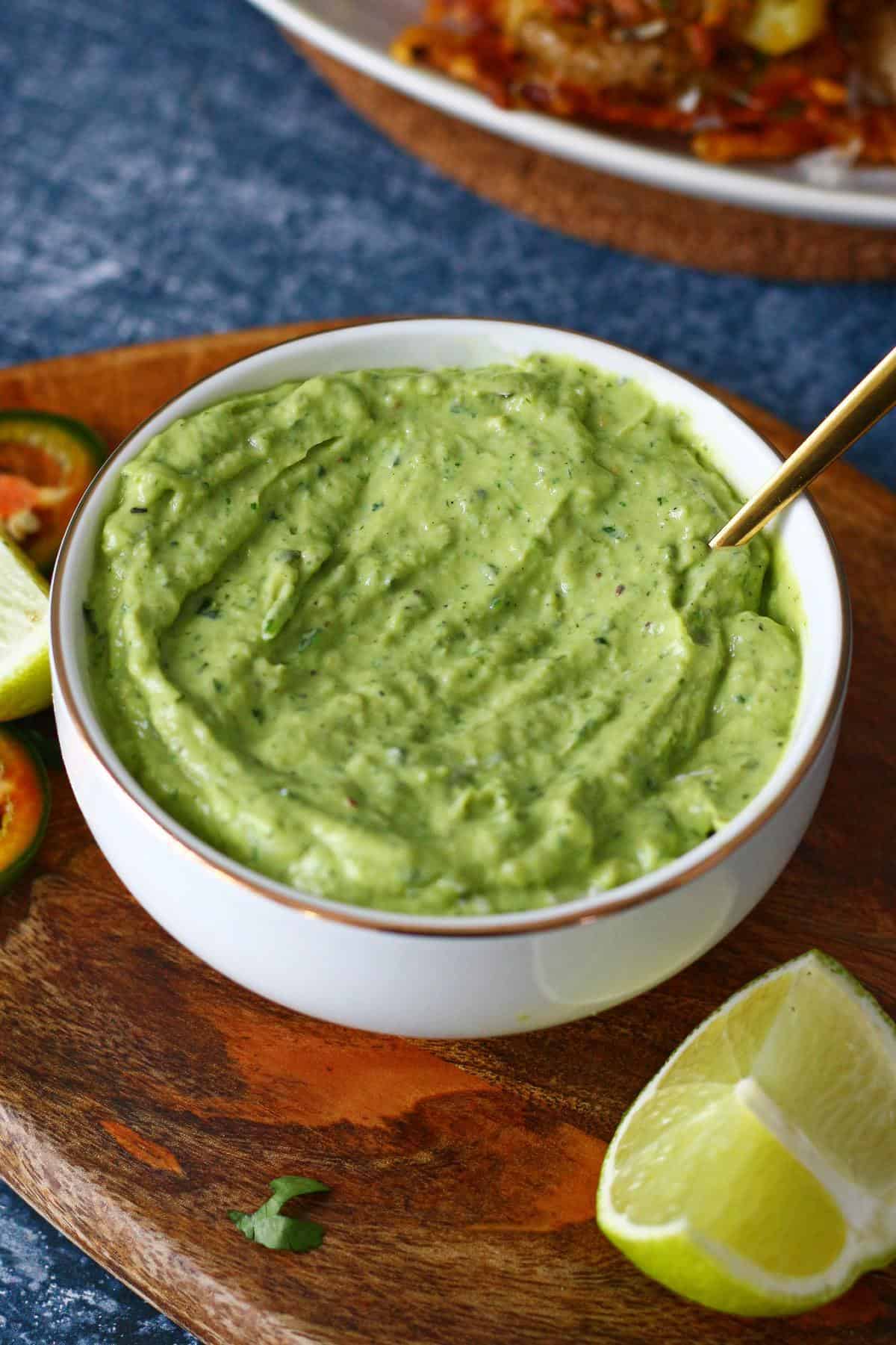 A bowl of avocado crema on a brown wooden board with lime wedge on the side.