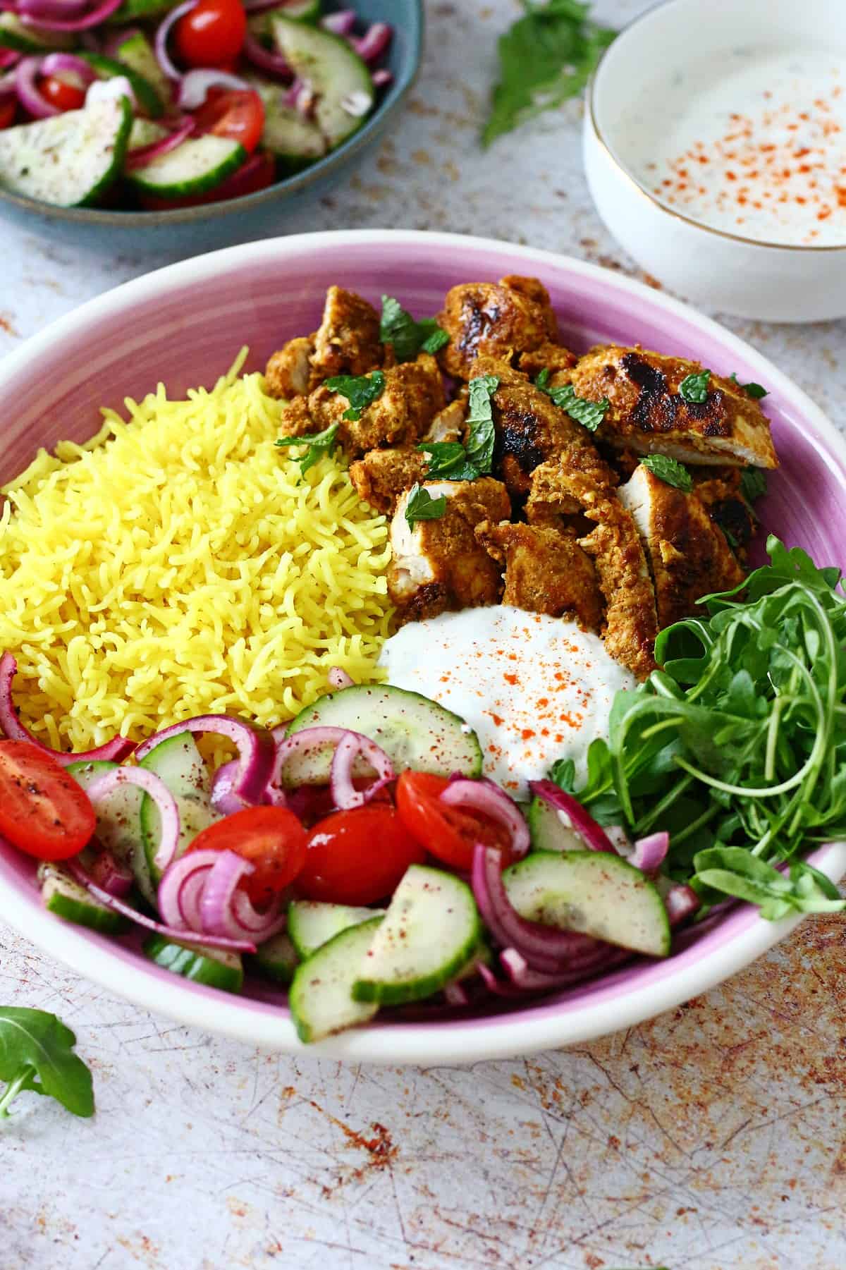 A shallow plate filled with yellow rice, grilled chicken strips, white sauce and salad with some sauce and salad at the back.