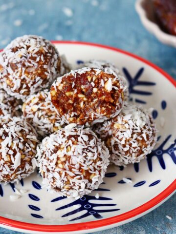 Coconut date balls on a plate with the top ball cut open on a blue backdrop.