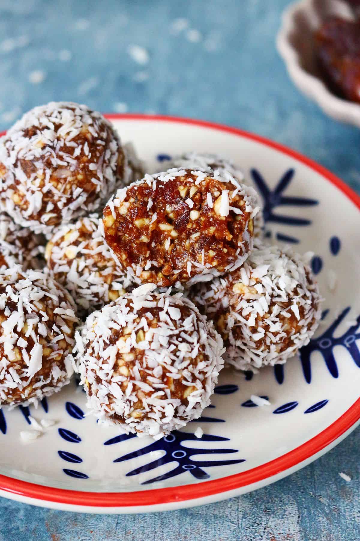 Coconut date balls on a plate with the top ball cut open on a blue backdrop.