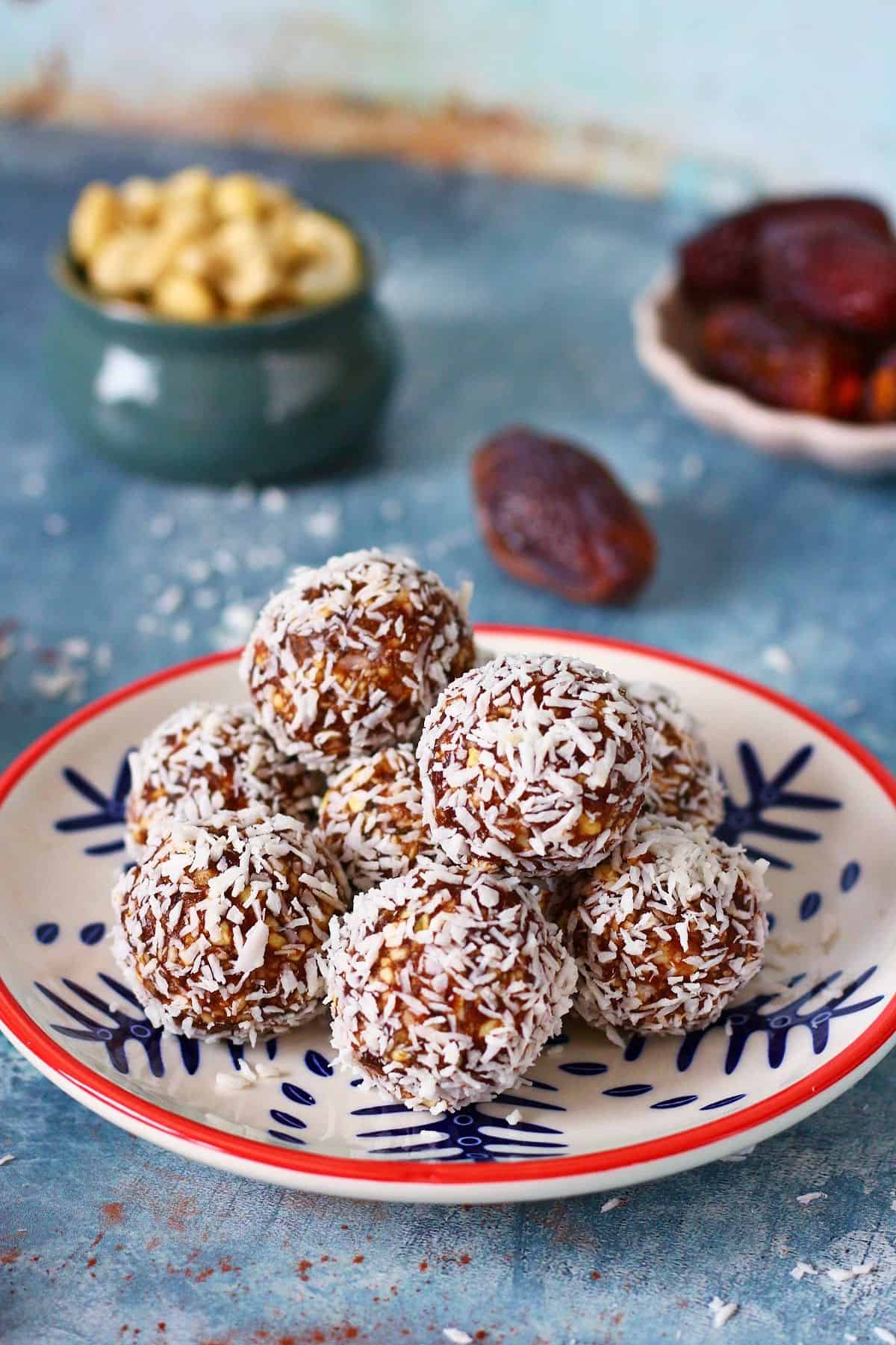 Coconut date balls stacked on a plate with whole dates and cashews behind.