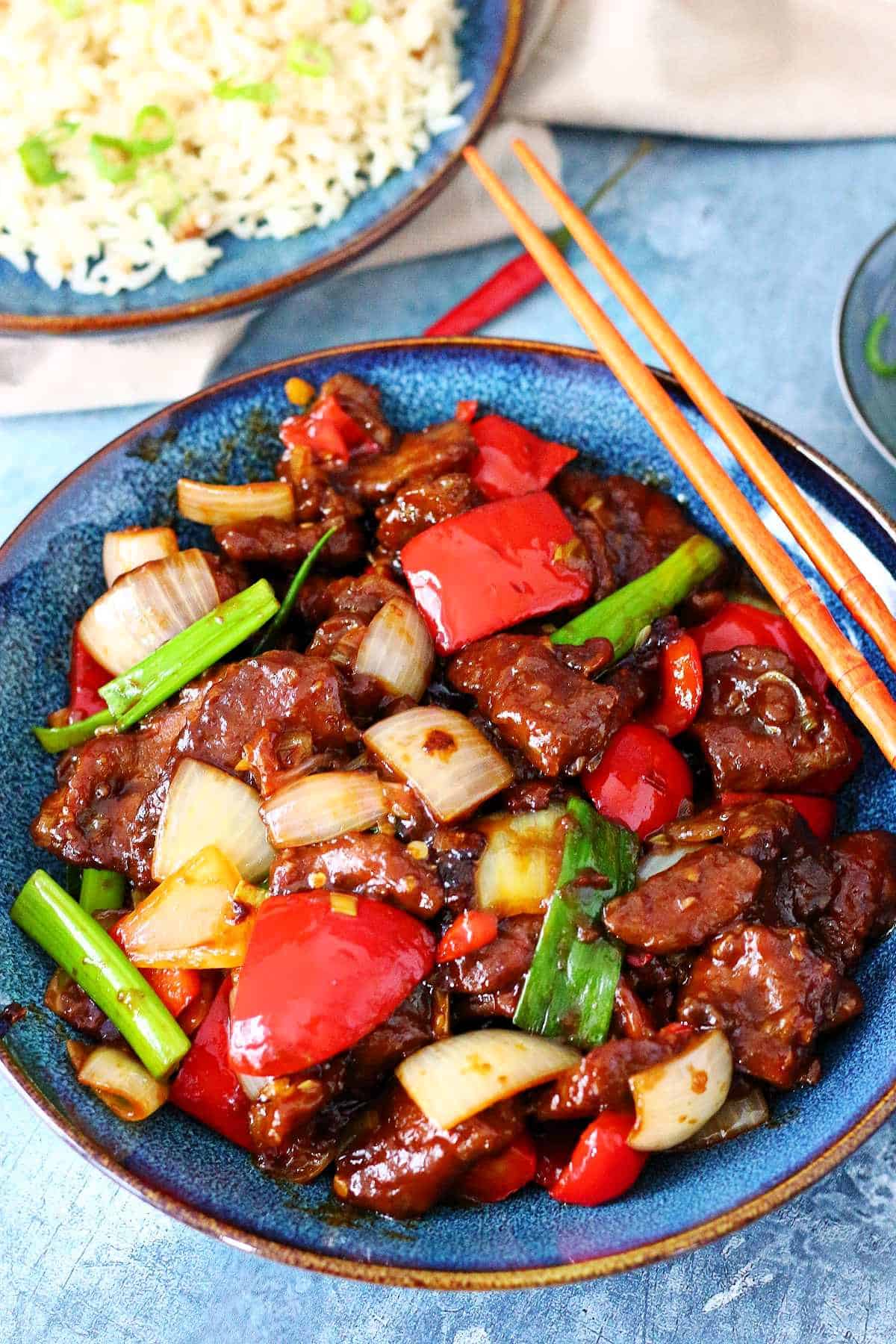 Beijing beef, cubed onions and red peppers in a blue plate with chopsticks on the side and rice at the back.