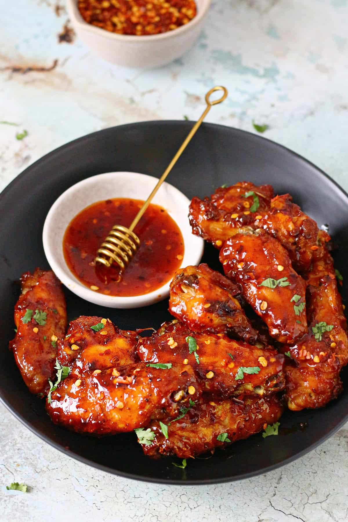 Sticky wings in a shallow plate with hot honey sauce at the side and red pepper flakes at the back.