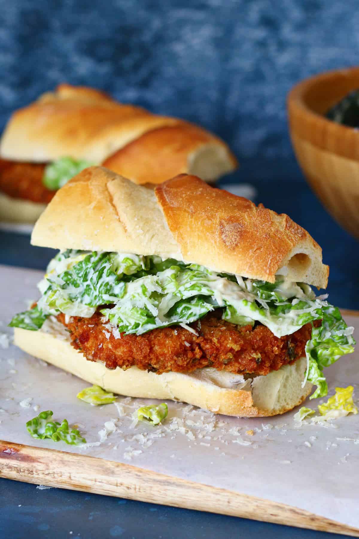 Chicken Caesar Sandwich on a white wooden board with butter paper and a sandwich at the back.