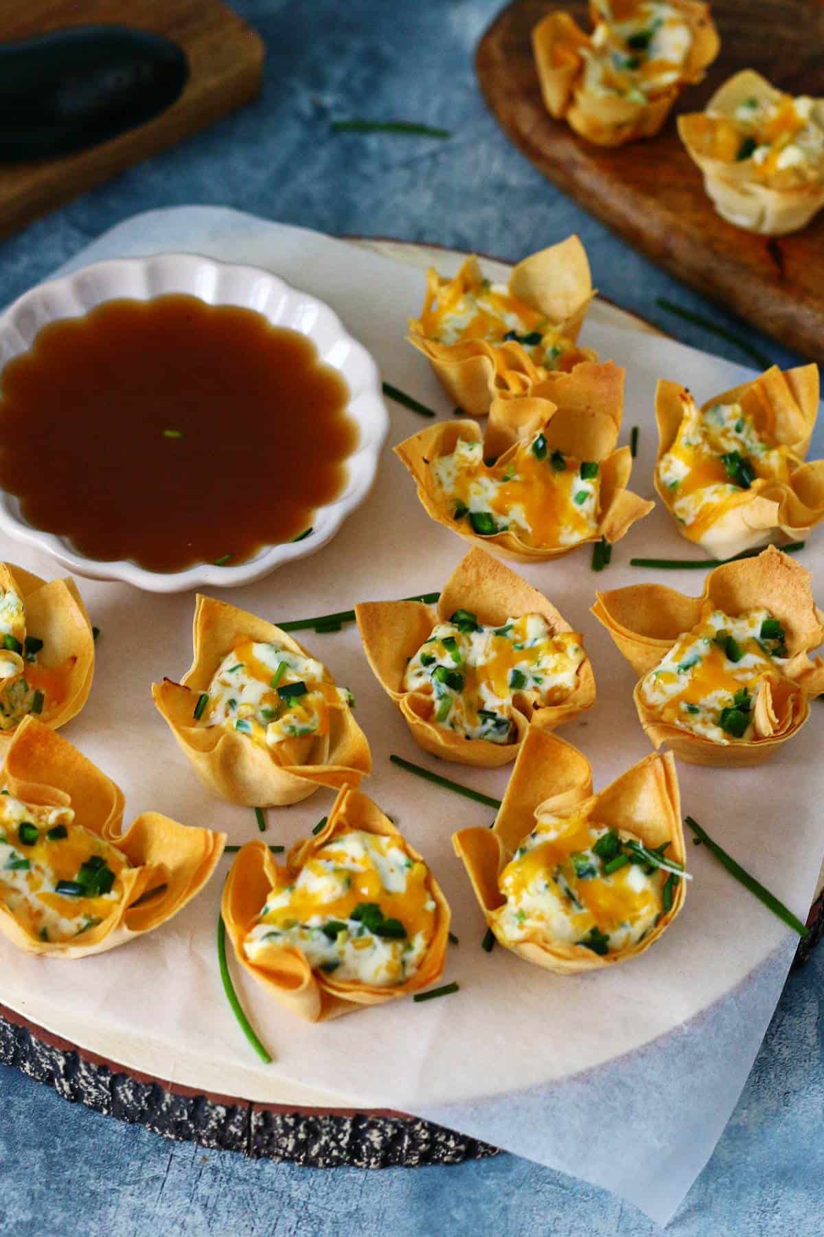 A dozen jalapeño popper wontons on a wooden serving board with a dip on the side.