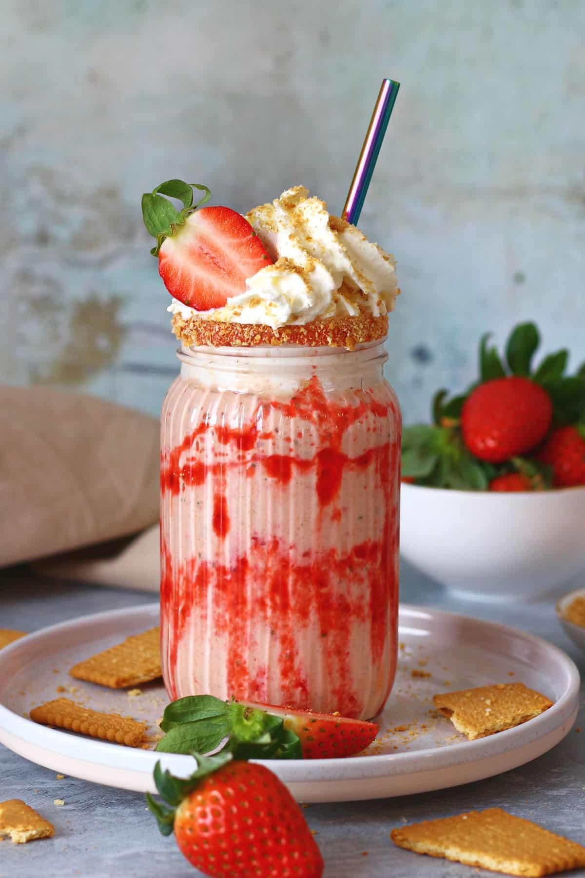 Strawberry cheesecake milkshake in a tall glass with toppings and straw surrounded by strawberries and broken crackers.