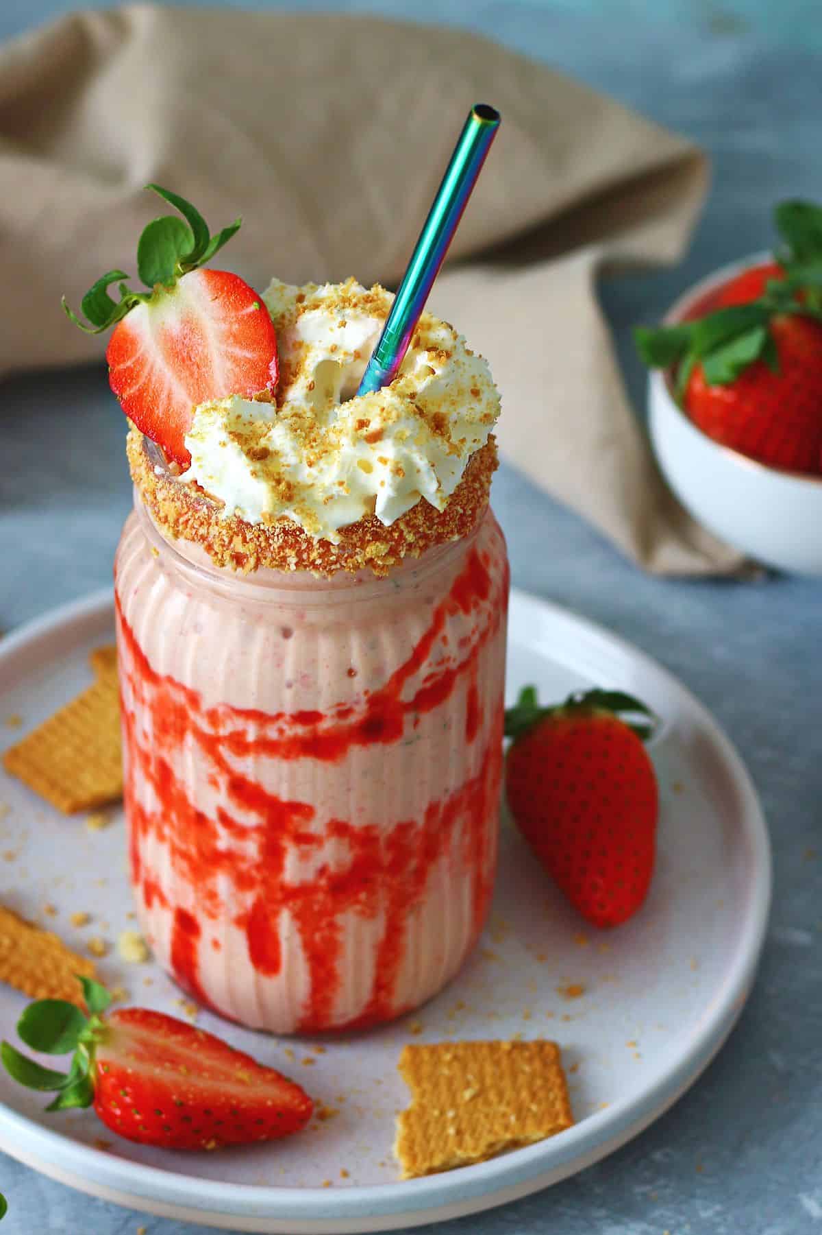 Strawberry cheesecake milkshake in a decorated glass topped with whipped cream and crushed graham crackers.