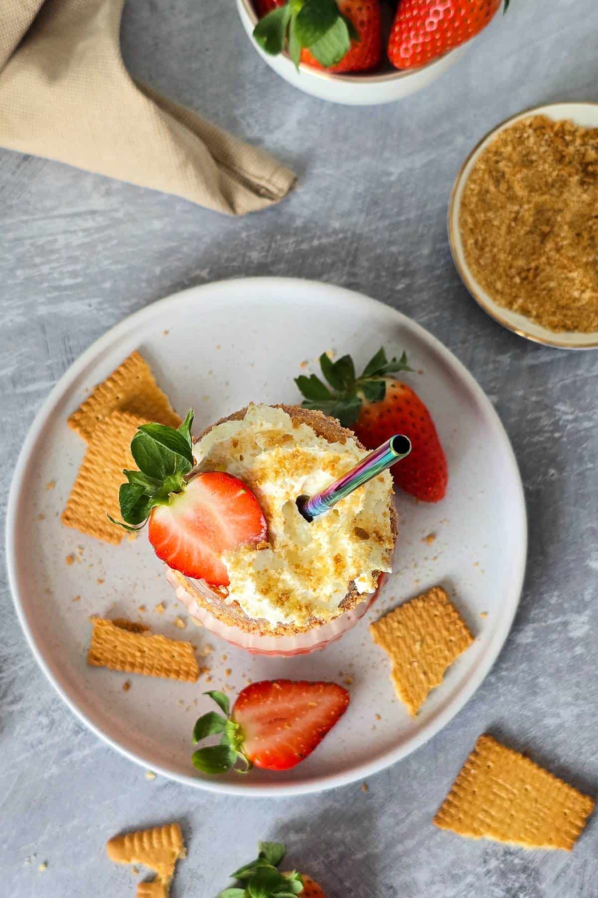 Overhead shot of strawberry cheesecake milkshake surrounded by fresh strawberries, broken and crushed crackers.