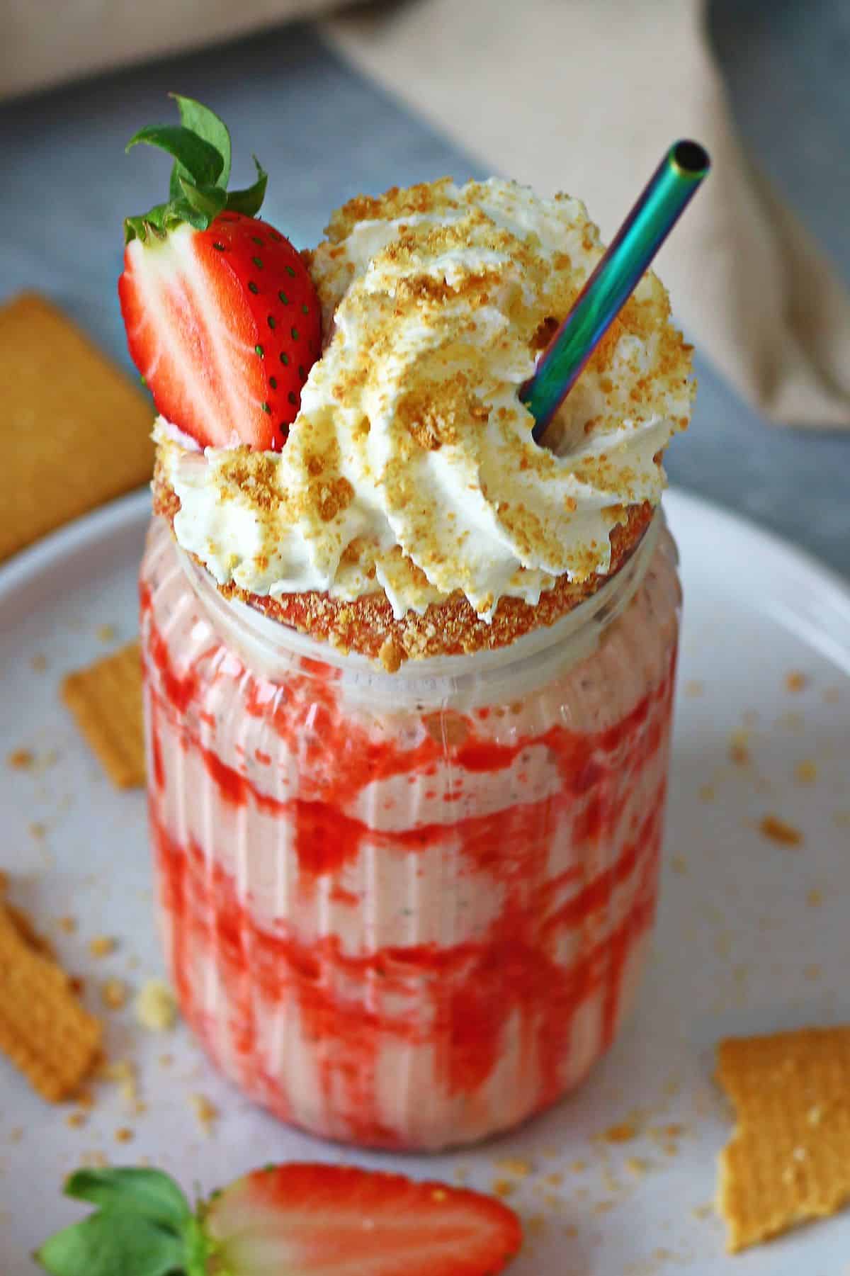 Strawberry milkshake with closeup toppings of whipped cream, sliced strawberry, crushed cookies and a metal straw.