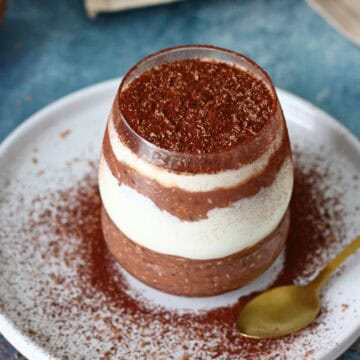 Tiramisu overnight oats in a glass on a white plate with gold spoon on the side.