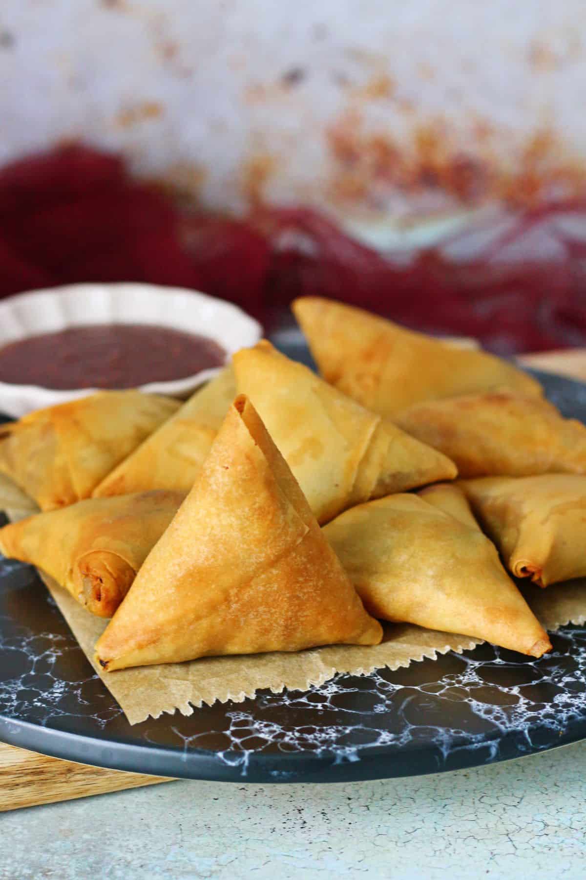 A group of keema samosas on a parchment paper on top of a black dish served with a side of ketchup.