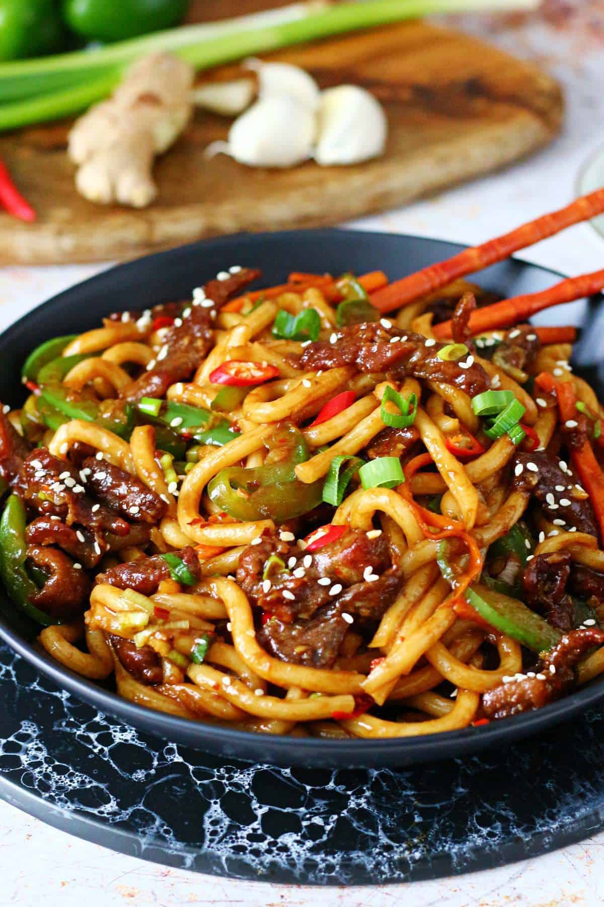 Mongolian beef noodles garnished with sesame seeds in a black dish with chopsticks, and vegetables behind it.