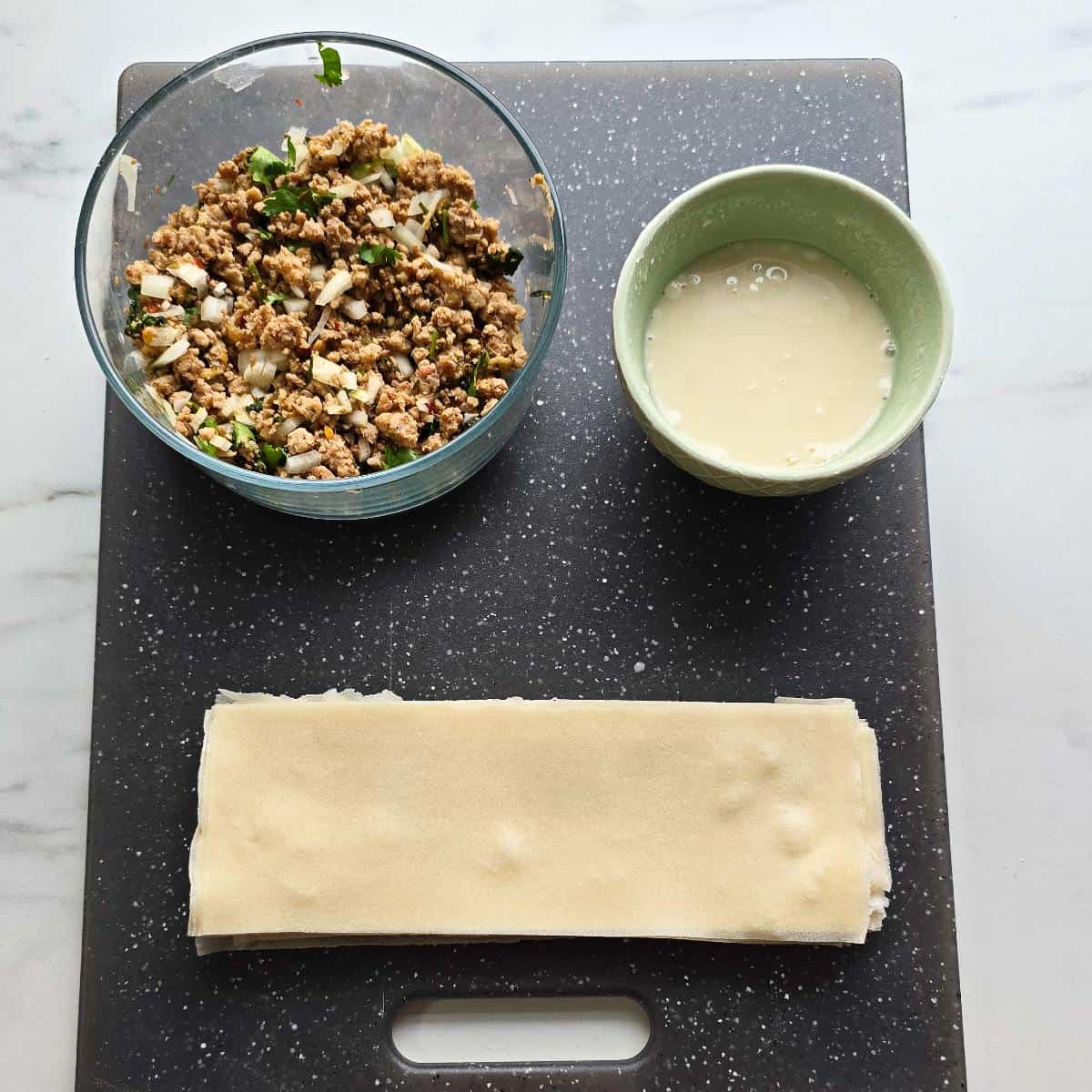 A chopping board with samosa sheets, meat mixture in a glass bowl and flour paste in a small bowl.